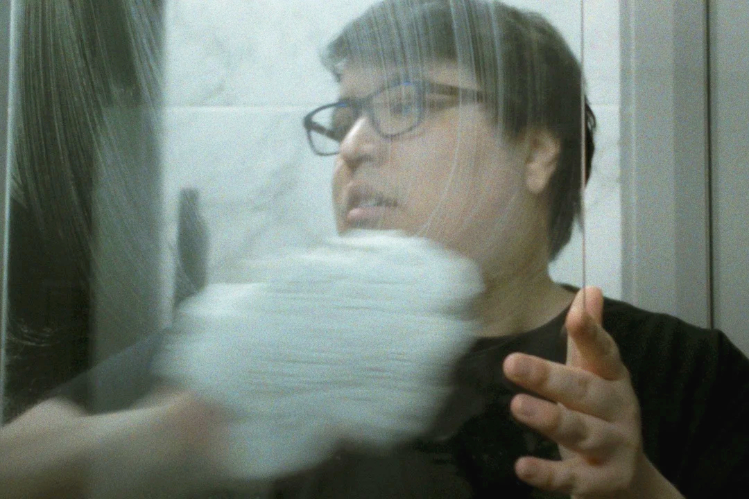 Still aus PAUL: Paul beim Putzen eines Badezimmers - schwarze Hornbrille, schwarzes Shirt, ein durchschnittlicher, geradezu langweiliger Kerl vom Äußeren her.