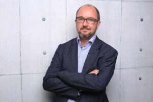 Der Redakteur Daniel Bax steht vor einer grauen Betonwand. Er trägt einen dunkelblauen Anzug, ein hellblaues Hemd und hat die Arme verschränkt.