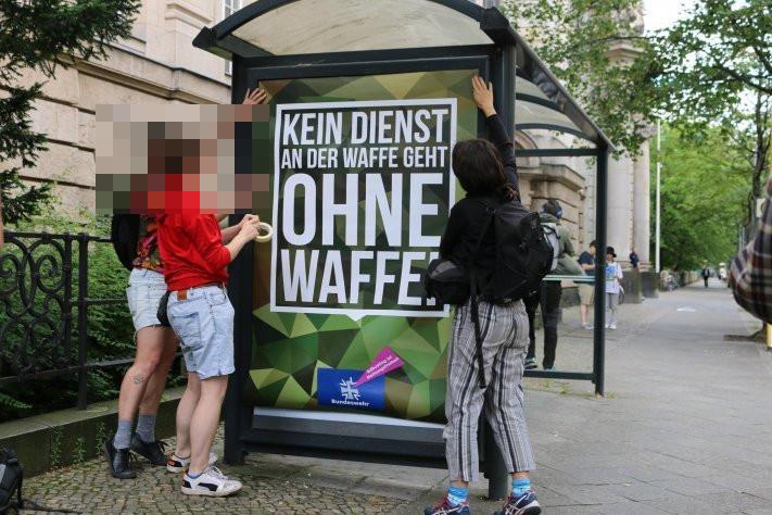 Auf diesem Foto sind drei Menschen zu sehen, die an der Werbevitrine einer Bushaltestelle mit einem vermeintlichen Werbeplakat der Bundeswehr zugange sind. Auf dem Plakat ist zu lesen: "Kein Dienst an der Waffe geht ohne Waffe!"