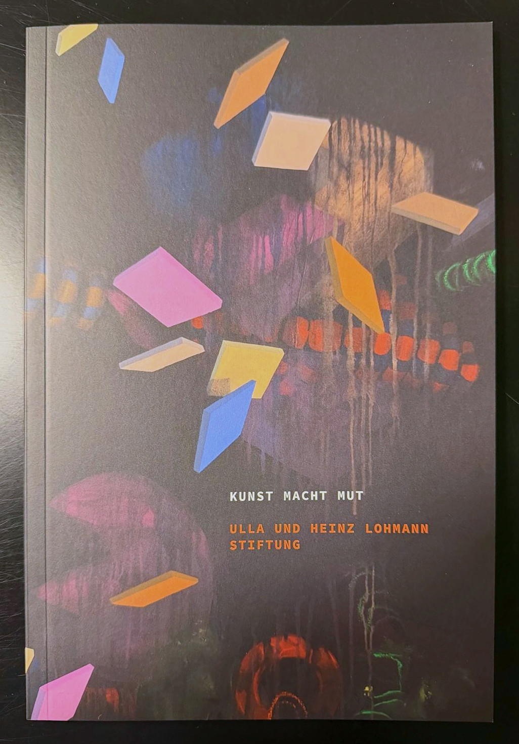 Titelseite des Katalogs &bdquo;Kunst macht Mut&ldquo; der Ulla und Heinz Lohmann Stiftung mit einem dunklen, gemalten Motiv mit schwebenden bunten Rauten in Rosa, Orange, Gelb und Blau.