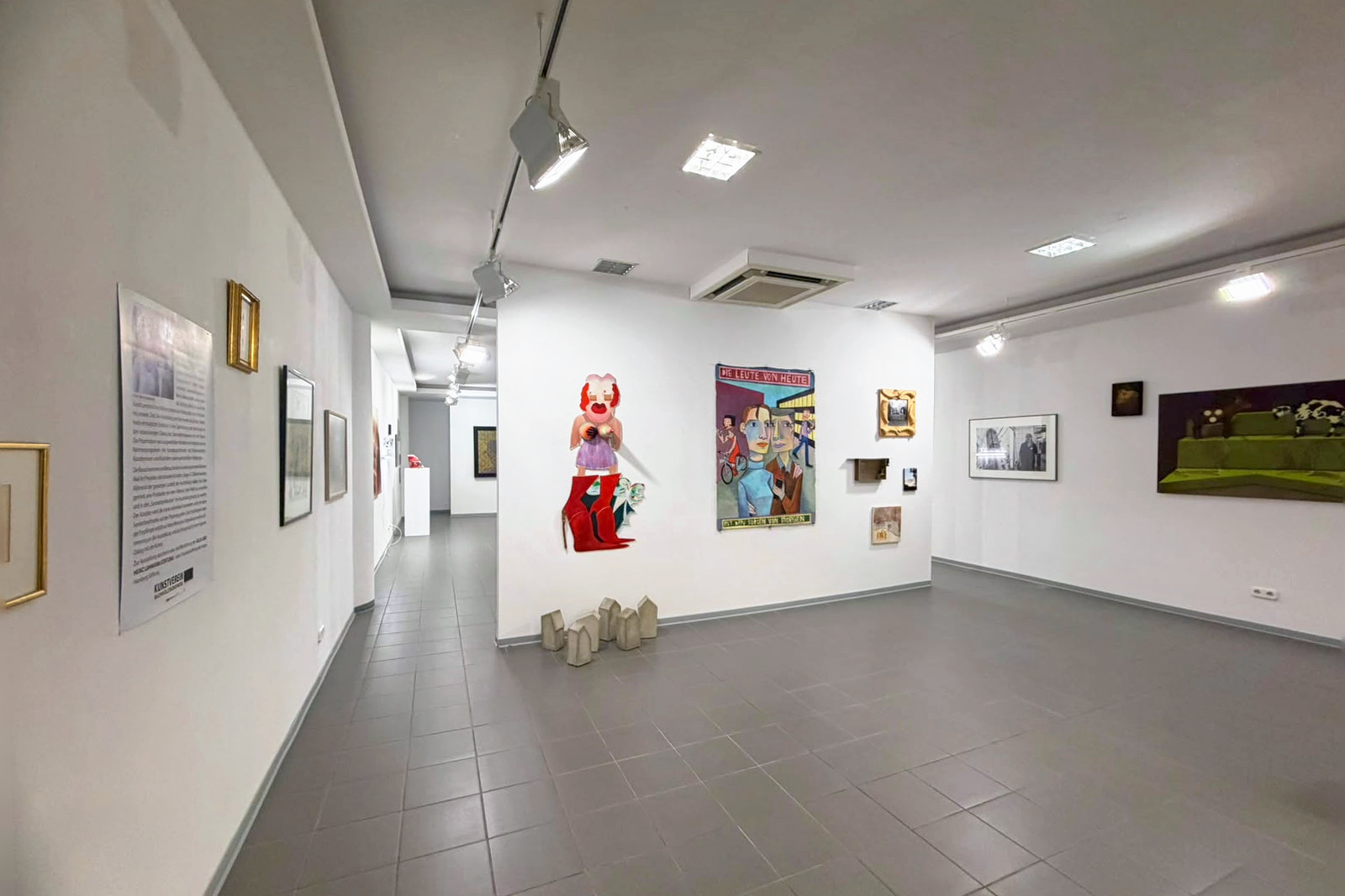 Ausstellungsraum mit wei&szlig;en W&auml;nden und grauem Fliesenboden. An einer Stellwand h&auml;ngen eine ausgeschnittene Figur mit roten Stiefeln und das Plakatbild &bdquo;Die Leute von heute&ldquo;, davor stehen kleine Betonh&auml;user auf dem Boden, rechts weitere Bilder.