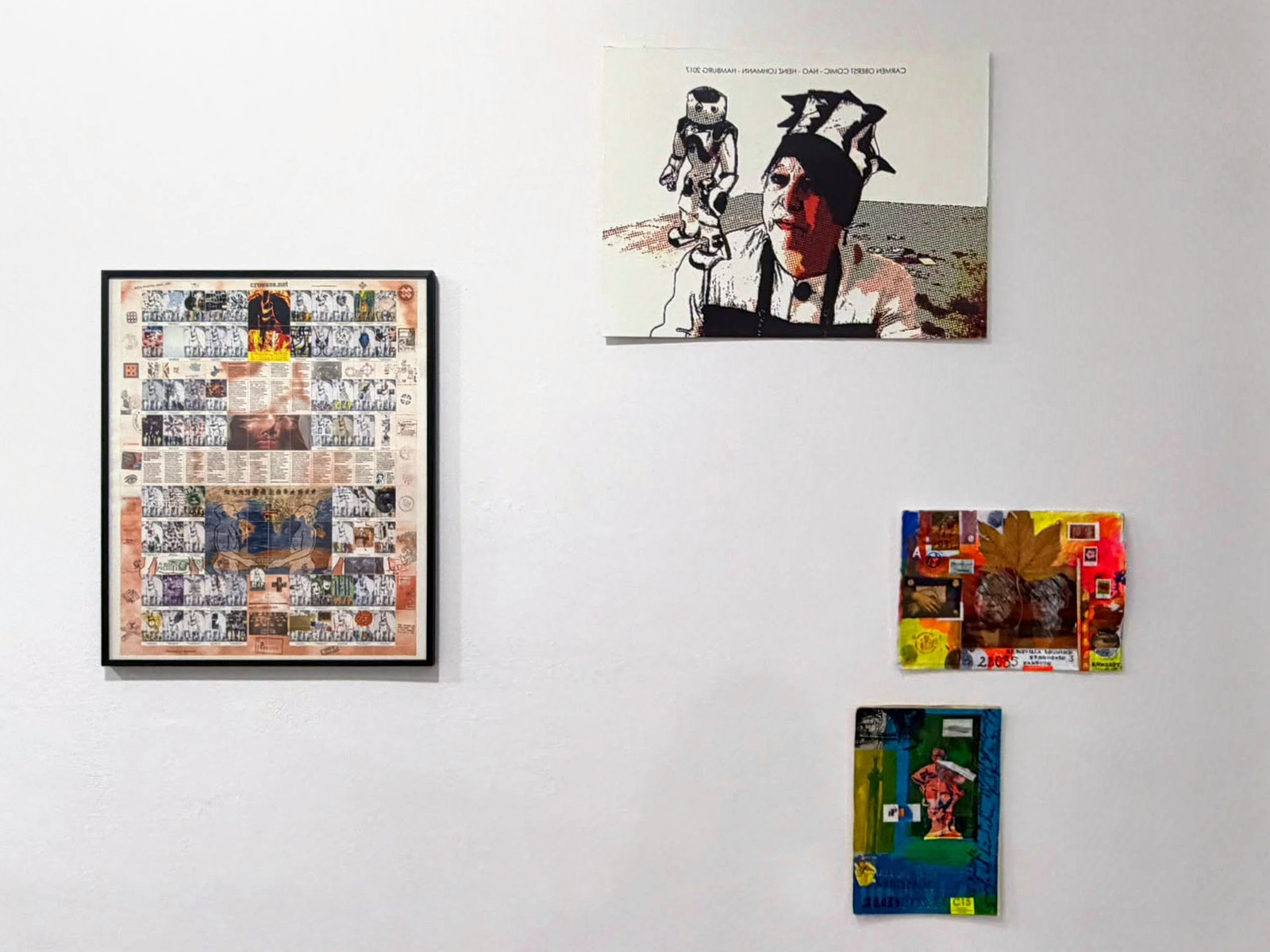 Wei&szlig;e Wand mit vier Arbeiten, links ein gerahmter Bogen voller kleiner K&uuml;nstlerbriefmarken mit einer Weltkarte in der Mitte, rechts oben ein Comic-Druck mit einem Roboter und einem Portr&auml;t, rechts unten zwei bunt gestaltete Mail-Art-Umschl&auml;ge.