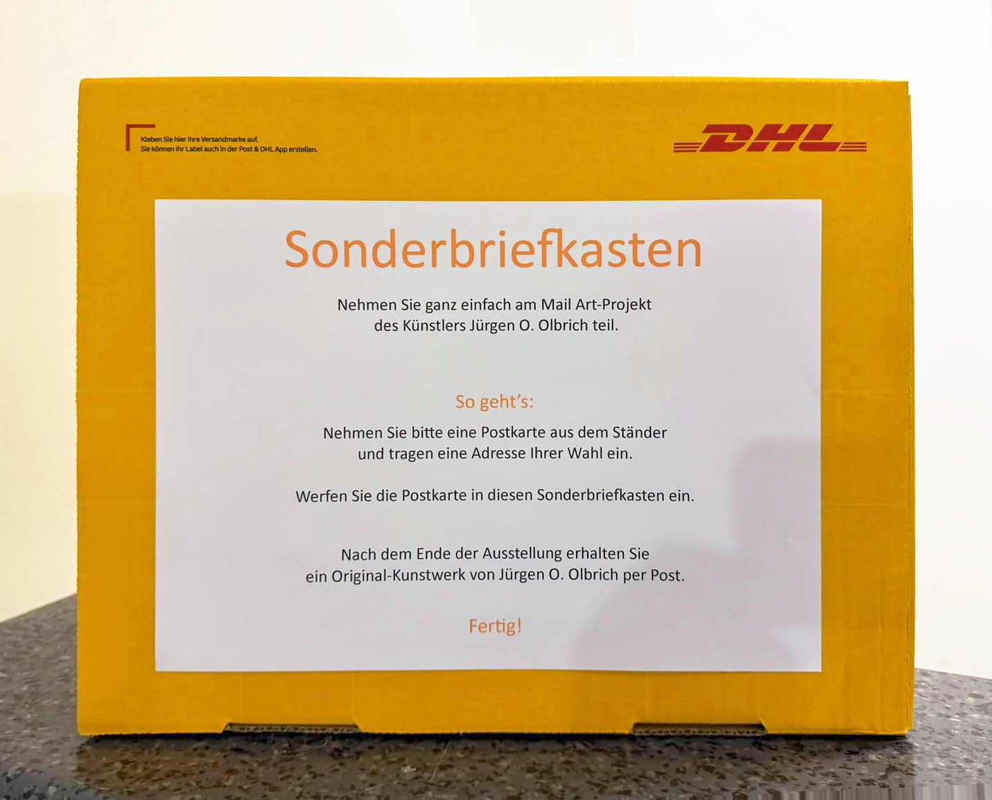 Gelber DHL-Karton als &bdquo;Sonderbriefkasten&ldquo; mit aufgeklebter Anleitung. Postkarte aus dem St&auml;nder nehmen, Adresse eintragen, einwerfen, und nach der Ausstellung kommt ein Original-Kunstwerk von J&uuml;rgen O. Olbrich per Post.